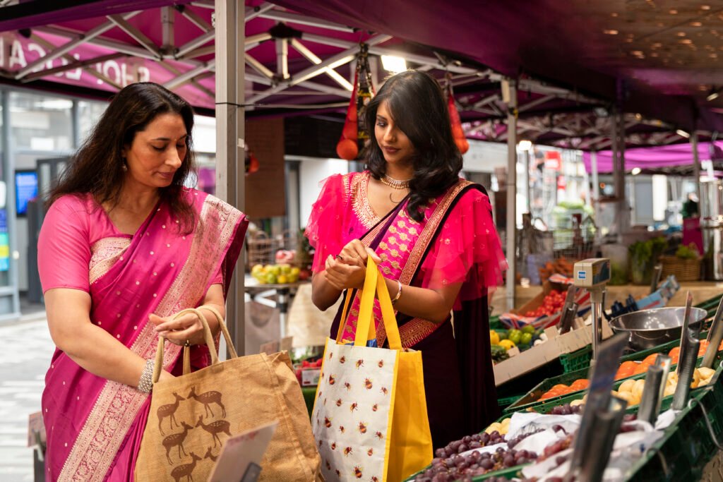 Two women in vibrant sarees shopping for fresh fruits and vegetables at an outdoor market, carrying reusable bags. Title: